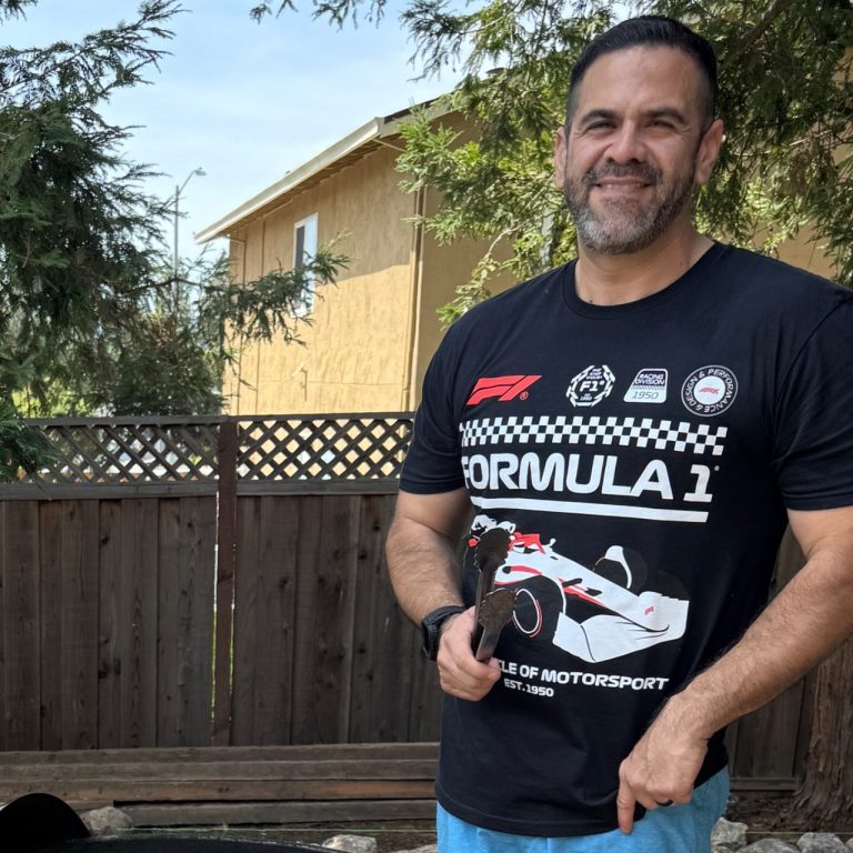 Man in a Formula 1 t-shirt stands outdoors near a wooden fence.
