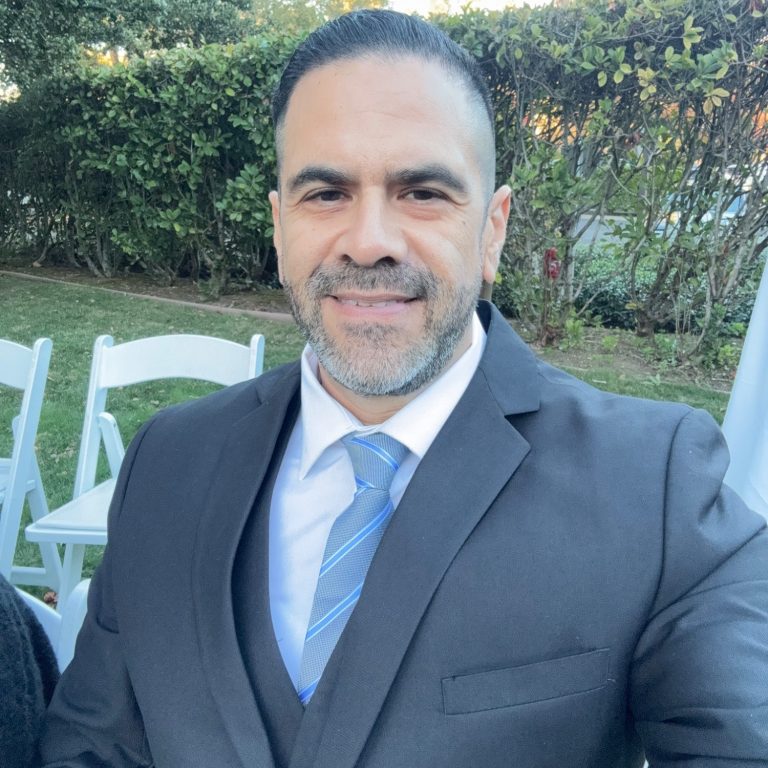Man in a suit and tie smiling outdoors, with chairs and greenery in the background.